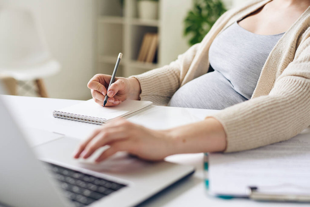 Крупним планом невизначена вагітна жінка сидить за столом і використовує ноутбук під час запису завдань у блокноті
 - Фото, зображення
