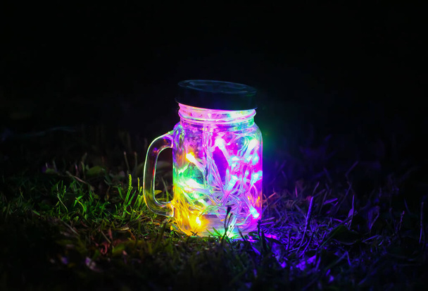 Святкова листівка з яскравими барвистими гірляндами у скляній банці на нічному фоні природи
. - Фото, зображення