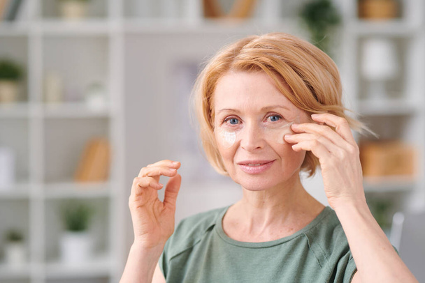 volwassen vrouw met blond haar zetten onder ooglapjes op haar gezicht terwijl staan in de voorkant van de camera tijdens schoonheid procedure thuis - Foto, afbeelding