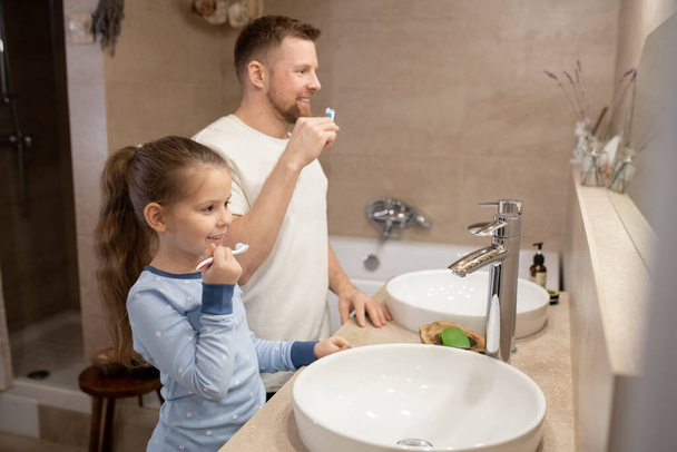 Linda niña y su feliz padre joven con cepillos de dientes va a cepillarse los dientes mientras está de pie en el baño delante del espejo
 - Foto, imagen
