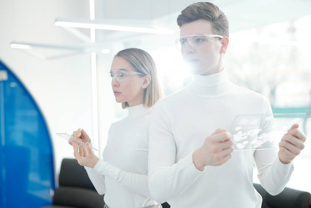 Dois jovens desenvolvedores de software progressivo contemporâneo em óculos de segurança e blusas brancas usando gadgets móveis futuristas durante o trabalho
 - Foto, Imagem