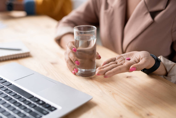 Close-up of unrecognizable businesswoman holding glass of water and taking pink pill at workplace - Foto, imagen