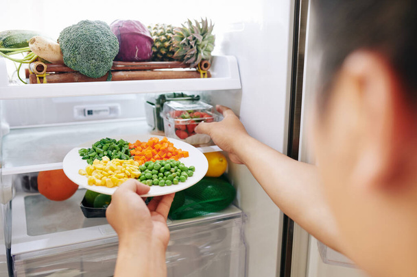 Женщина берет тарелку с кукурузой, горохом, морковью и зелеными бобами и контейнер с клубникой из холодильника
 - Фото, изображение