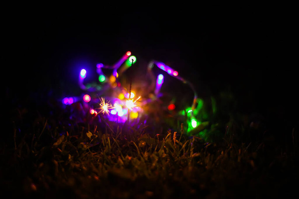 Biglietto festivo con brillanti luci colorate ghirlanda e fuoco bengalese in vaso di vetro su sfondo natura notte. - Foto, immagini
