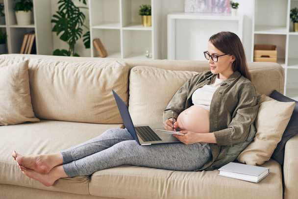 Atractiva joven embarazada en ropa de casa sentada en el sofá y leyendo el artículo en el ordenador portátil mientras toma notas sobre la maternidad
 - Foto, Imagen