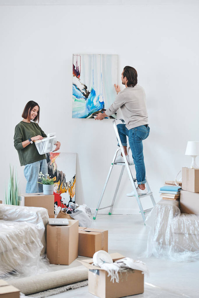 Νεαρός άνδρας με casualwear στέκεται στη σκάλα από τοίχο σε τοίχο και κρέμεται αφηρημένη ζωγραφική, ενώ η σύζυγός του κοιτάζοντας την εικόνα στο πλαίσιο - Φωτογραφία, εικόνα