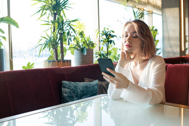 Hezké mladé blond žena v bílé hedvábné halence rolování v smartphonu, zatímco sedí u stolu na gauči v pohodlné restauraci - Fotografie, Obrázek