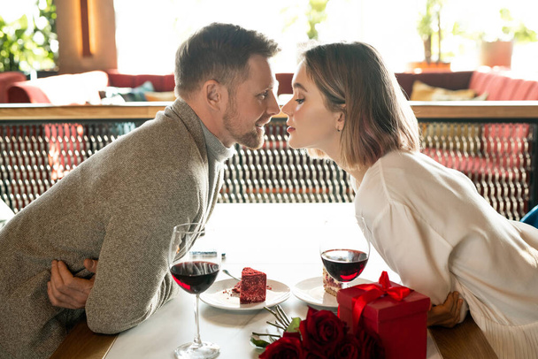 Gelukkig jong aanhankelijk paar buigen over serveertafel, terwijl het houden van hun gezichten dicht bij elkaar tijdens romantische date in restaurant - Foto, afbeelding