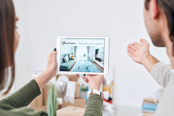 Mujer joven sosteniendo touchpad con la publicidad de nuevos bienes raíces en la pantalla, mientras que su marido expresando su opinión sobre su interior
 - Foto, imagen
