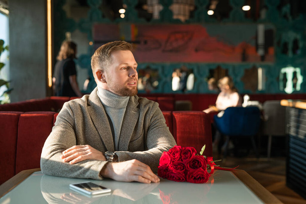 Joven con ramo o rosas rojas y teléfono inteligente sentado a la mesa en un lujoso restaurante mientras espera a su novia
 - Foto, Imagen
