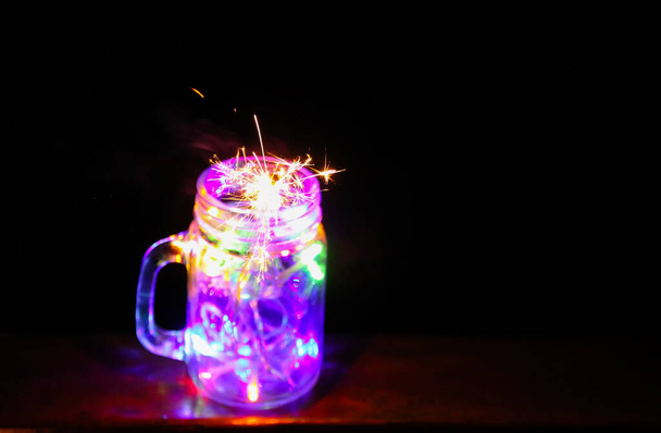 Ünnepi kártya fényes színes koszorúslámpák és bengáli tűz üveg üveg az éjszakai természet háttér. - Fotó, kép