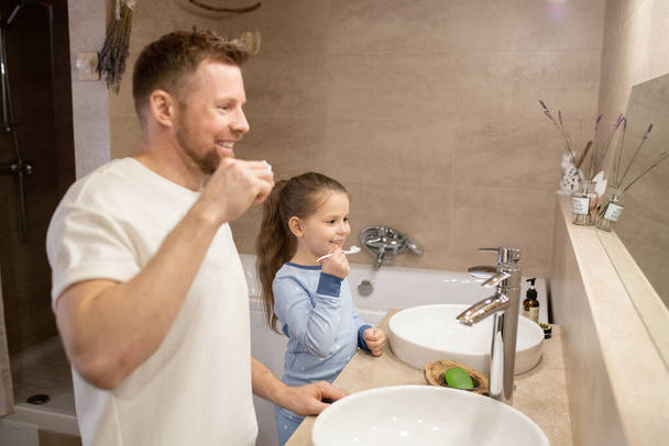 Feliz jovem pai barbudo e sua filha alegre bonito segurando escovas de dentes por seus dentes enquanto em pé no banheiro
 - Foto, Imagem