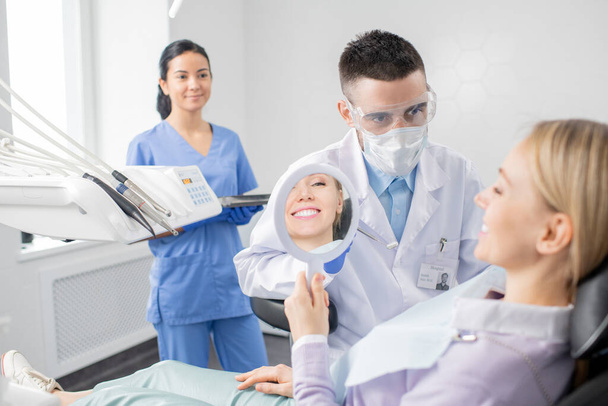 Счастливая молодая пациентка с зубастой улыбкой, смотрящая в зеркало, сидя в кресле в кабинете стоматолога со своим стоматологом впереди
 - Фото, изображение