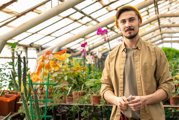Portrait of content hipster bearded nursery worker in cap holding trowel in greenhouse with flowers - Foto, imagen