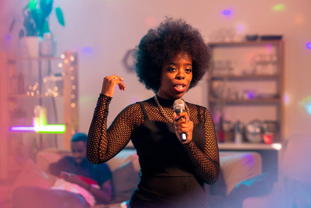 Молодая элегантная женщина в элегантном черном платье поет караоке в микрофон на фоне друзей на диване на домашней вечеринке - Фото, изображение