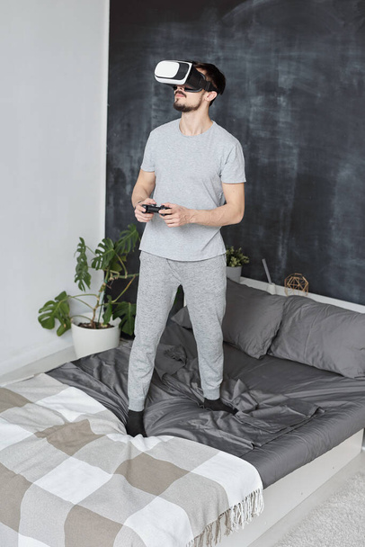 Contenuto bel giovane uomo con la barba in piedi sul letto e utilizzando joystick durante la riproduzione di videogiochi in occhiali di realtà virtuale
 - Foto, immagini