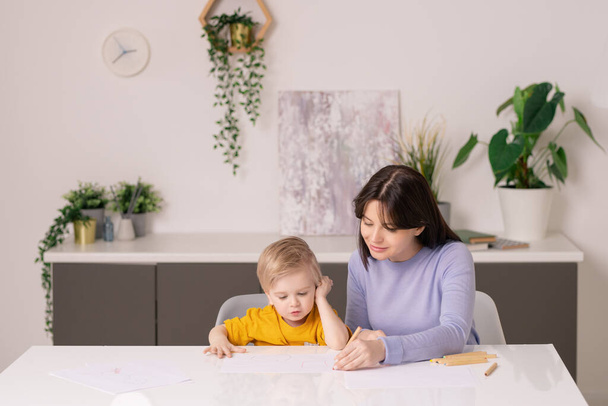 Young casual woman helping her cute little son to draw picture on paper with colorful crayons while both sitting by table in living-room - Photo, Image
