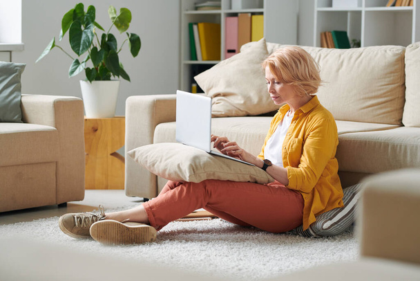 Mujer de negocios madura seria sentada en el suelo por el sofá con el ordenador portátil en la almohada en las rodillas y el trabajo de planificación en el entorno del hogar
 - Foto, Imagen