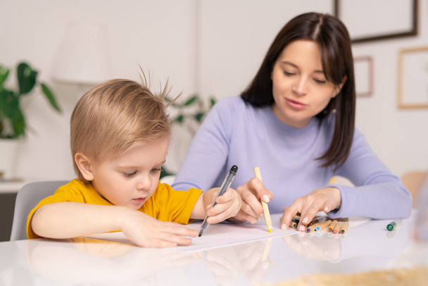 Vorsichtige junge Mutter sitzt mit kleinem Sohn am Tisch und zeichnet auf Papier - Foto, Bild
