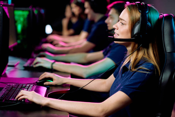 Girl in headset with microphone sitting at table and playing computer game in e-sport club - Foto, Imagen