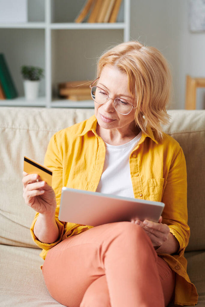 Блондинка зрелая женщина с тачпадом и кредитной картой сидя на диване после рабочего дня и покупки через интернет в гостиной
 - Фото, изображение