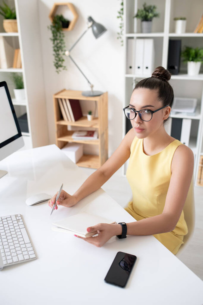 Portret van een serieuze jonge brunette zakenvrouw in gele jurk zit aan het bureau en het maken van notities in het dagboek tijdens het plannen van het werk activiteit - Foto, afbeelding