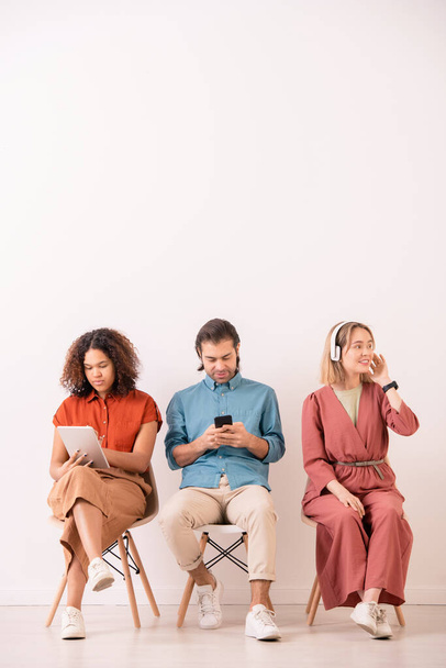 Young multi-ethnic man and woman interacting on social media using gadgets while positive girl listening to music in headphones - Foto, afbeelding