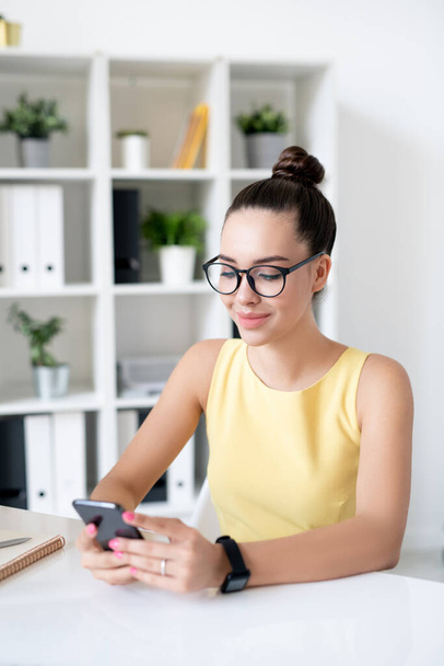 Usmívající se atraktivní mladá podnikatelka s vlasy houska sedí u stolu a pomocí smartphone při surfování sociálních médií během přestávky - Fotografie, Obrázek