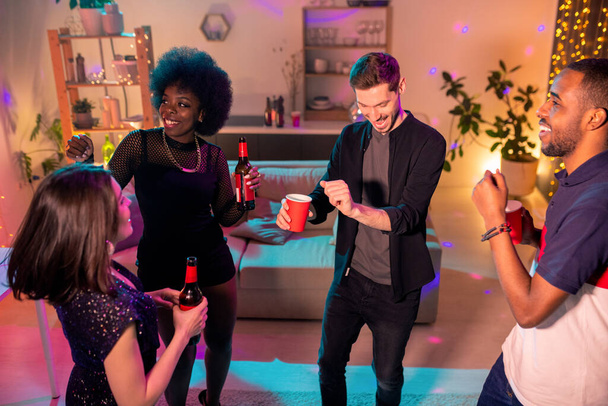 Молодые экстатические друзья, танцующие дома в гостиной и пивающие пиво на фоне дивана - Фото, изображение