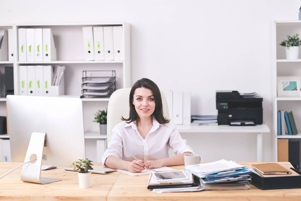 Jeune femme d'affaires prospère en tenue de cérémonie assise au bureau, faisant un plan de travail ou des notes sur un nouveau projet d'entreprise
 - Photo, image