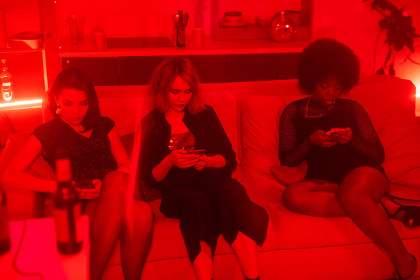 Rij van drie interculturele meisjes in glamoureuze jurken scrollen in smartphones terwijl ontspannen op de bank thuis partij - Foto, afbeelding