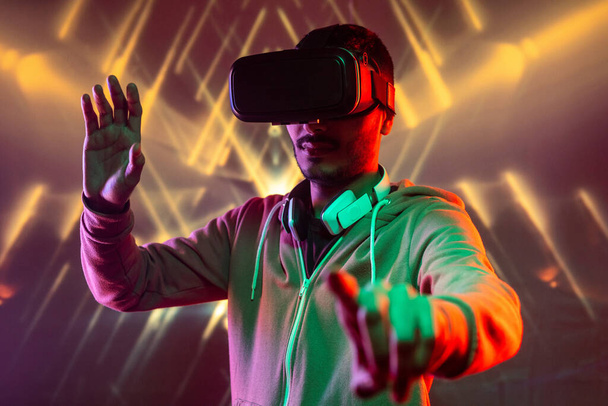 Jeune homme avec casque autour du cou gesticulant les mains tout en obtenant une expérience de réalité virtuelle sur fond géométrique lumineux
 - Photo, image
