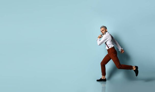 Gray-haired, bearded male in white shirt, brown pants and suspenders, black loafers. He is running against blue background - Φωτογραφία, εικόνα