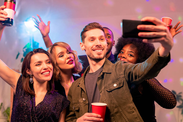 Akıllı telefonlu mutlu genç adam ve üç kültürler arası neşeli kız selfie çekiyorlar ve ev partisinde içkilerle neşeleniyorlar. - Fotoğraf, Görsel