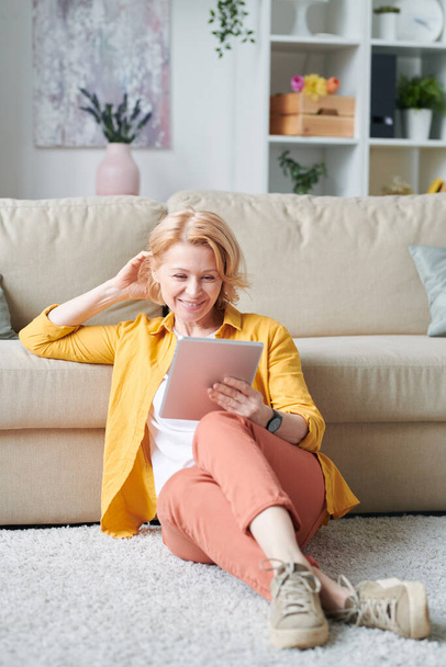 Wesoły dojrzewanie kobieta w casualwear patrząc na tablet wyświetlacz podczas oglądania online wideo na podłodze przez kanapę w środowisku domowym - Zdjęcie, obraz