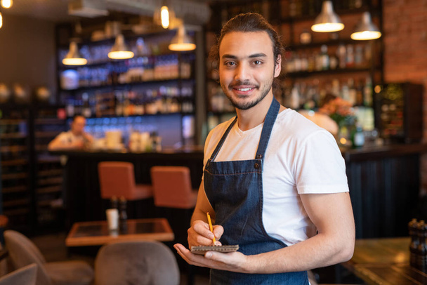 Young smiling waiter in workwear making notes in notepad while standing in front of camera on background of bar counter and interior - Photo, Image