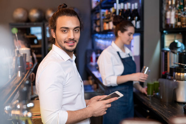 Jeune serveur amical en chemise blanche défilant dans le menu en ligne tout en vérifiant les prix des boissons et de la nourriture sur fond de collègue
 - Photo, image