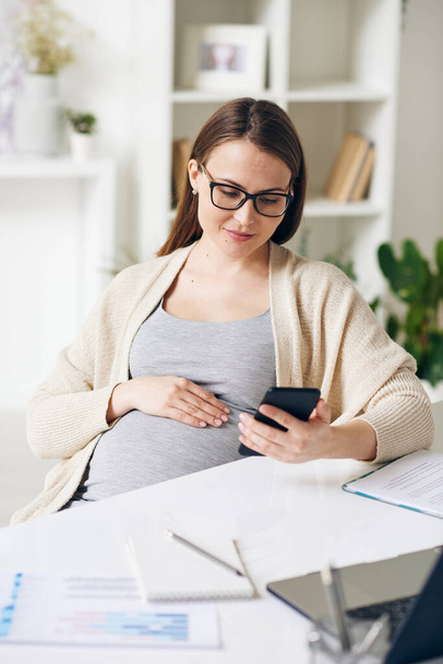 Giovane donna d'affari incinta in occhiali seduti alla scrivania e controllo messaggero telefonico in ufficio
 - Foto, immagini