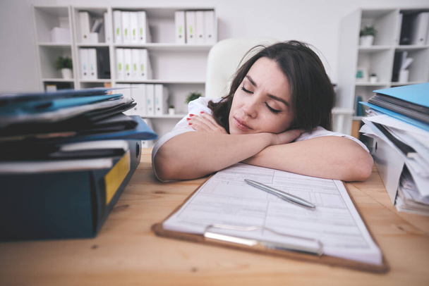 Mujer joven y cansada gerente de oficina o contable durmiendo en el escritorio frente al portapapeles con papeles financieros después de un duro día de trabajo
 - Foto, imagen