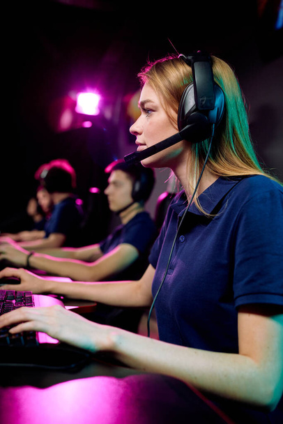 Zaměřeno mladá žena počítačový hráč v hands-free sluchátka s mikrofonem pomocí klávesnice při hraní online hry v e-sportu klubu - Fotografie, Obrázek