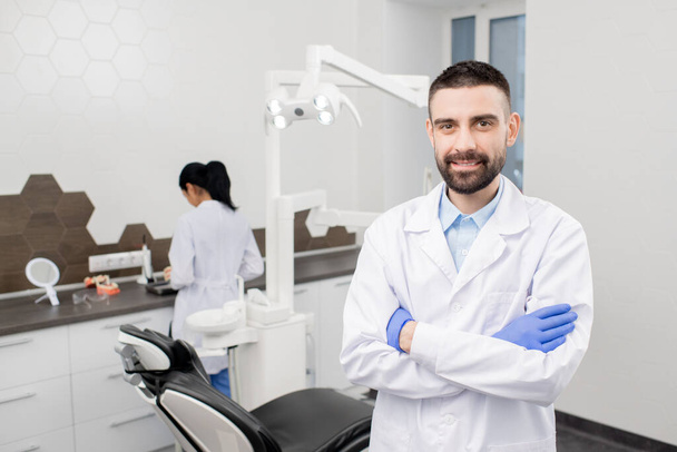 Szczęśliwy młody brodaty profesjonalny dentysta w rękawiczkach i białym płaszczu stojący przed kamerą na tle asystenta - Zdjęcie, obraz