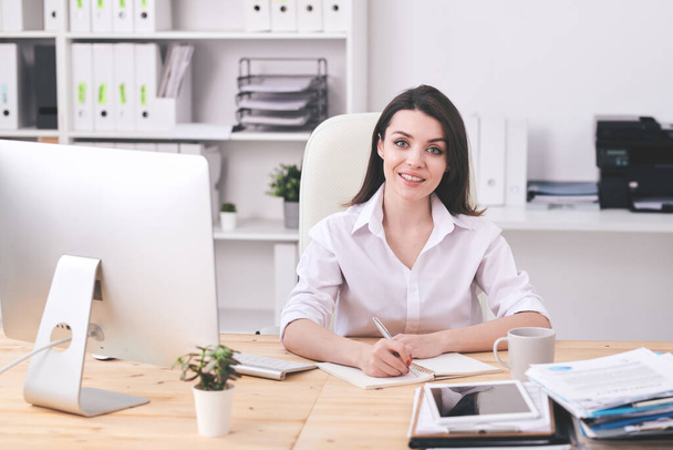 Счастливая молодая брюнетка деловая женщина в формальной одежде глядя на вас, записывая план рабочего дня утром в офисе
 - Фото, изображение