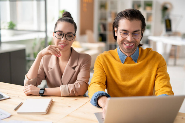 Pozytywni młodzi współpracownicy biznesowi w okularach siedzący przy stole i korzystający z laptopa podczas pracy nad projektem internetowym - Zdjęcie, obraz