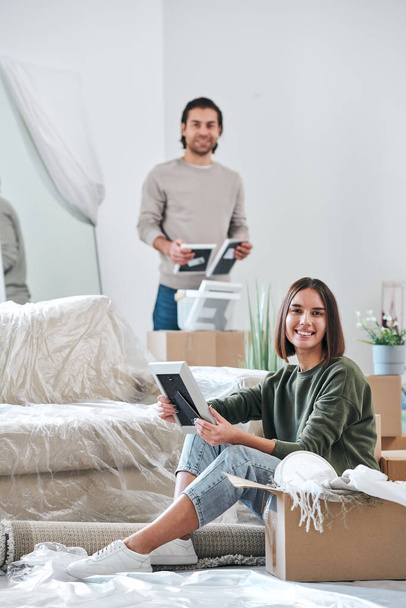 Young smiling female with picture frame sitting on the floor and looking at you while her husband unpacking boxes with stuff on background - Photo, Image