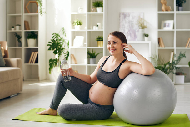 Smiling healthy young pregnant woman in sportswear sitting on yoga mat and leaning on fitball while drinking water at home - Foto, Imagen