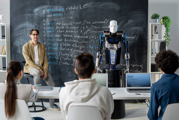 Grupo de estudantes adolescentes olhando para o robô de pé ao lado de quadro negro e descrevendo suas características técnicas para o professor no seminário - Foto, Imagem