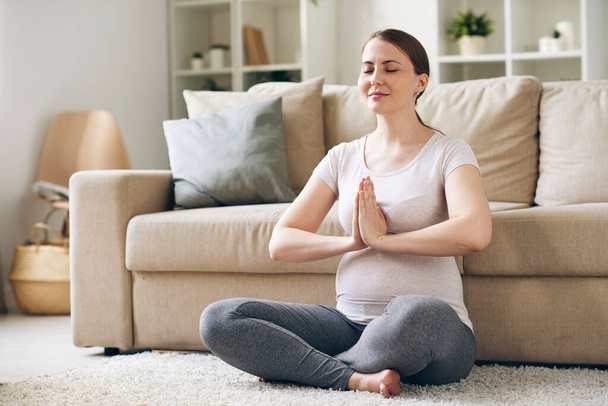 zadowolona młoda kobieta w ciąży siedzi z rękami w Namaste i medytuje z zamkniętymi oczami w domu - Zdjęcie, obraz