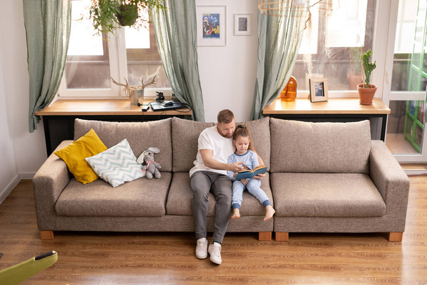 Junger Vater und seine süße kleine Tochter sitzen auf der Couch und lesen Geschichten oder Märchen, während sie zu Hause in Quarantäne bleiben - Foto, Bild