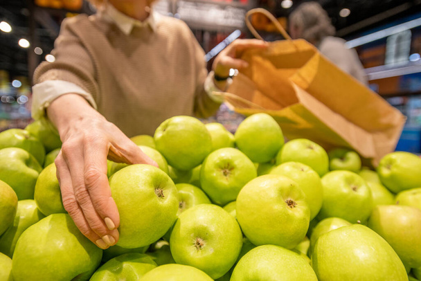 Käsi vuotiaiden naisten kanssa paperipussi ottaen tuoretta mummo seppä omena seistessään hedelmien näyttö nykyajan supermarketissa
 - Valokuva, kuva