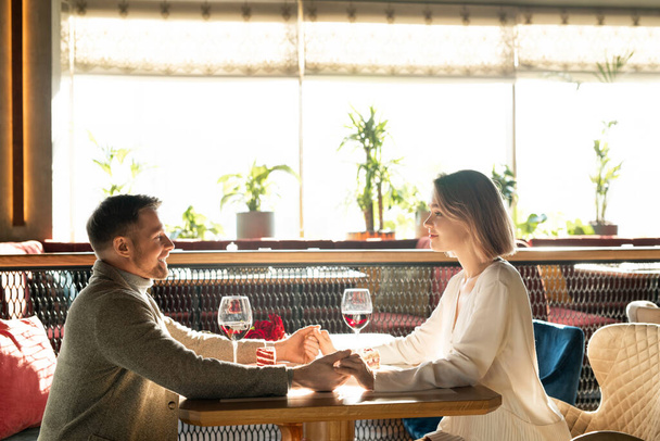 mooi jong vrouw en haar vriend zitten door geserveerd tafel voor elkaar en vasthouden door handen tijdens romantische date - Foto, afbeelding